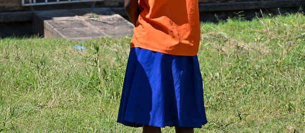 Back view of a child in an orange shirt and blue shorts standing on grass near a brick building.