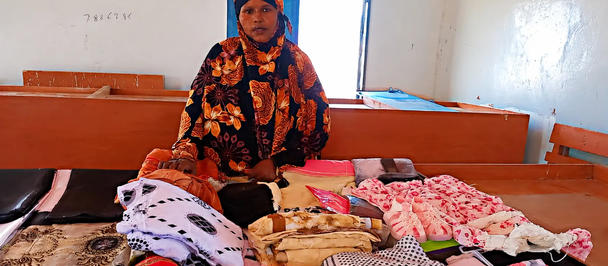 A woman in a colorful floral hijab stands beside a bed covered with folded clothes in a small room.