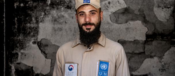Bearded man in beige uniform with logo patches and cap, standing against a textured wall.