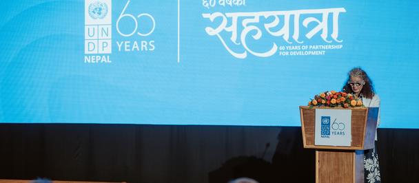 Photograph: woman speaks at a podium on a bright blue stage with UNDP Nepal 60th logo.