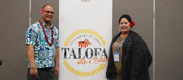 Two adults with lanyards pose beside a banner with a yellow laurel circle in a conference room.