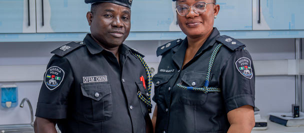 Two police officers in dark uniforms with berets stand side by side in a lab with blue cabinets.