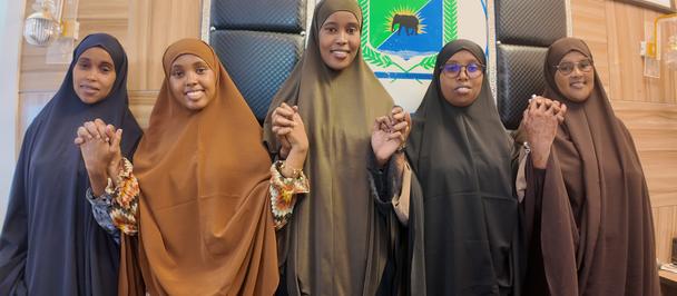 Six women in colorful hijabs and abayas stand in a row on steps in front of a yellow building.