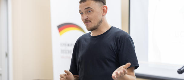 A man speaks during an event at the veterans’ centre in Ripky, Chernihiv Oblast, gesturing with his hands while standing in front of a presentation screen. A banner acknowledging the support of the Government of Germany is visible in the background.