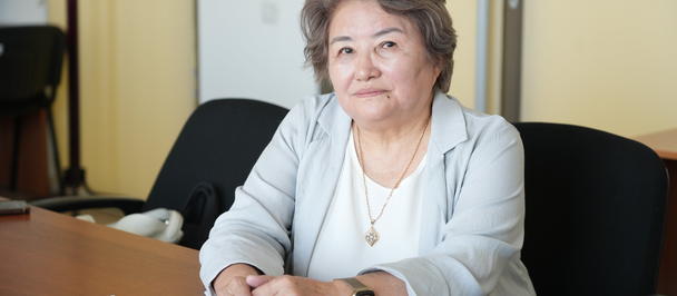 Older woman with gray hair sits at a wooden desk in an office, hands folded, gentle smile.