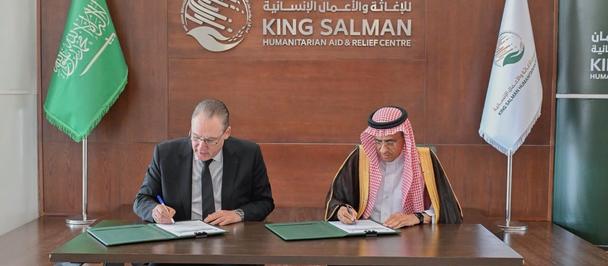 Two men signing documents at a formal ceremony, with a King Salman backdrop and flags.