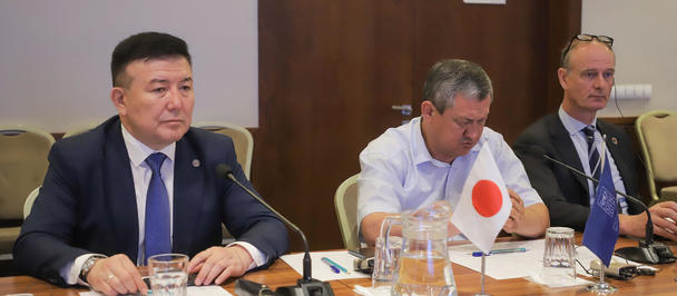 Three men in suits sit at a conference table with microphones and papers.
