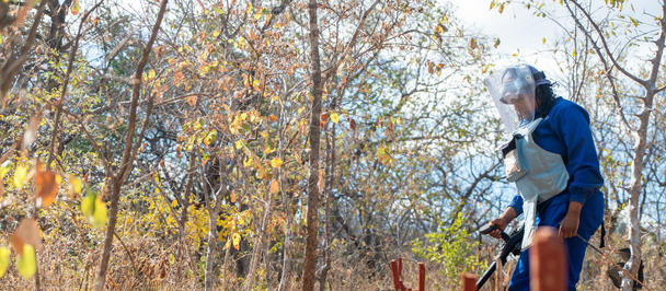 Person in blue coveralls and gloves standing among autumn leaves, working on a fence post.