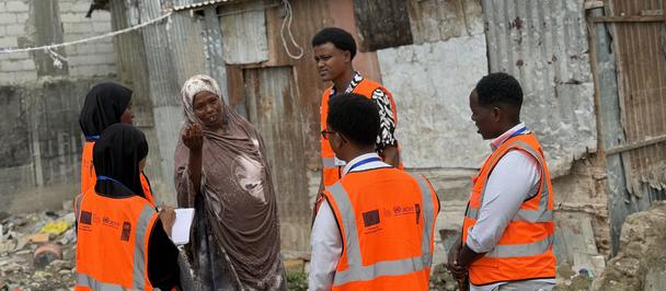 Group of people in orange safety vests talking near rundown wooden shacks