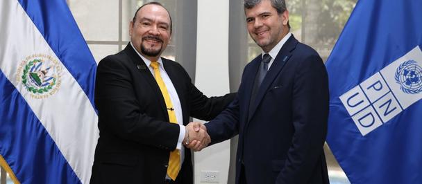 Two men in suits shake hands, smiling, in front of El Salvador and UNIDO flags.