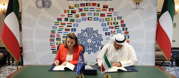 Photo: Two officials sign documents at a table, Kuwaiti flags on both sides, colorful backdrop.