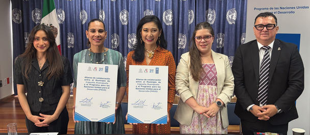 Five people in a formal meeting room; two women hold documents; flags and blue backdrop.