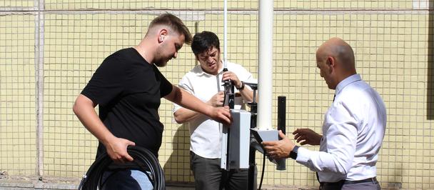 Three men inspect a post-mounted device beside a chain-link fence.