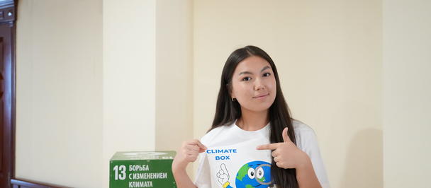 Girl in a white T-shirt with a globe logo holds a note, standing beside a green recycling sign.