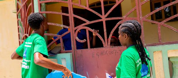 Photo of two students in green shirts beside a pink metal gate, one holding a blue folder.