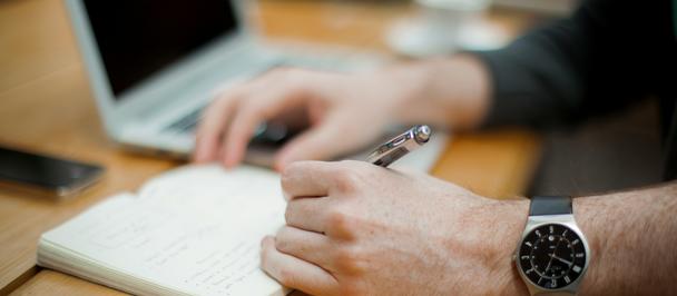 Close-up of hands with a pen writing in a notebook on a wooden desk, laptop nearby.