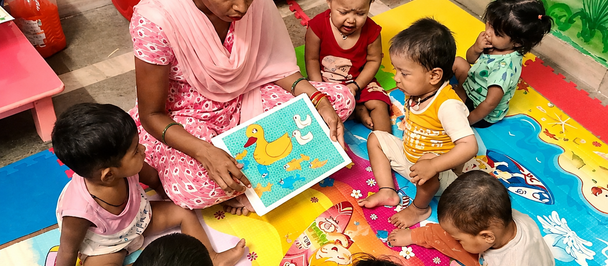 Photograph of a teacher reading a colorful book to kids on a bright classroom rug.