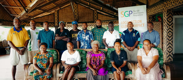 Group of diverse adults posing in a thatch-roofed hall; banner with a logo in the back.
