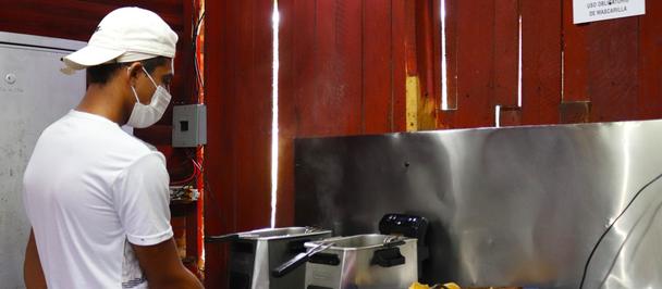 Cook in white shirt and cap at a stainless steel kitchen station with a blue bin.
