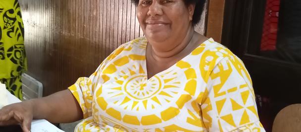 Smiling Black woman in a yellow and white patterned dress sits at a table indoors.