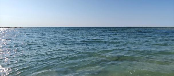 Photo: Calm coastal scene with turquoise water, sunlit rocks near shore, and a clear blue sky.