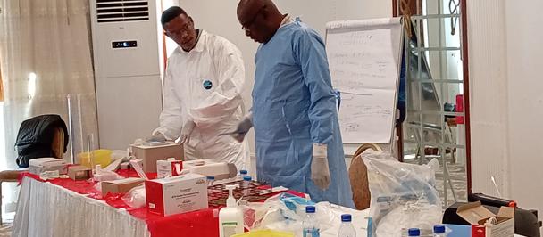 Photo: two researchers in lab coats stand by a long table with supplies in a clinical room.