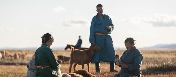 Blue-robed man on a hay bale; two men sit in a rural field with a brown animal nearby.