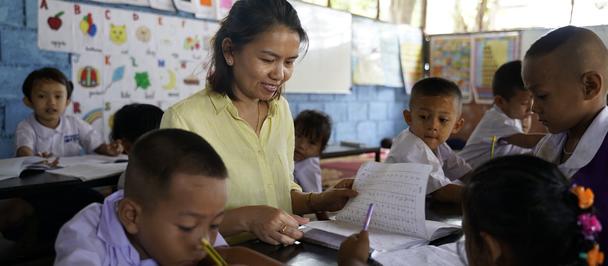 Photograph: teacher assists students at desks in a bright classroom with colorful posters.