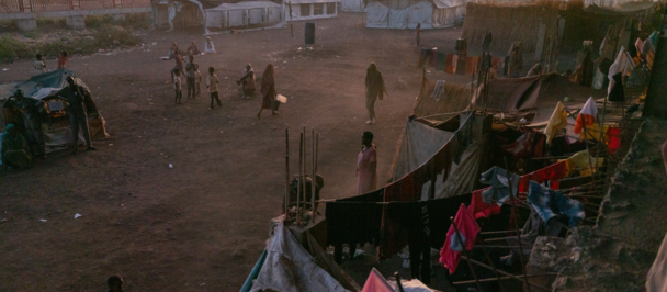 Desert campsite at dusk with tents, orange sky, people milling about, and stalls to the right.