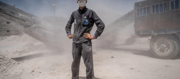 Dusty construction site with worker in hard hat and mask, hands on hips; truck in background.