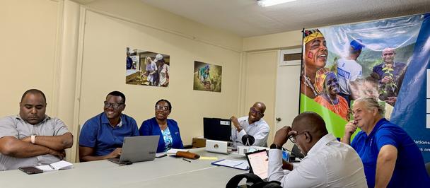 People sit around a rectangular table in a beige meeting room with posters and a colorful banner.