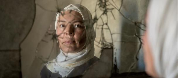Bandaged woman reflected in a cracked mirror; out-of-focus figure in the foreground.