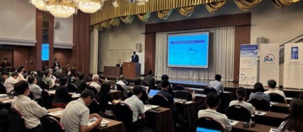 Conference room filled with attendees watching a speaker at a podium; presentation on a screen.