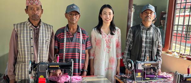 Four individuals stand behind sewing tables with machines and fabric in a bright room.