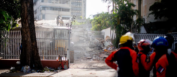 Photograph: dusty urban street with debris from earthquakes and barricade; rescue team in hard hats and orange vests.
