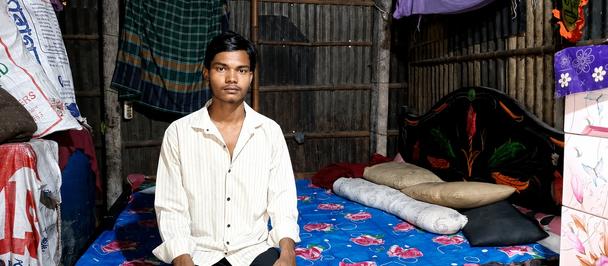 Man in a white shirt sits on a blue-patterned bed inside a rustic bamboo hut.