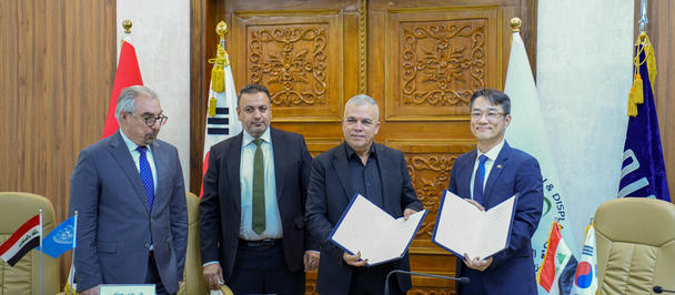 Photograph of five men in suits signing documents at a formal ceremony, with flags and a crest.