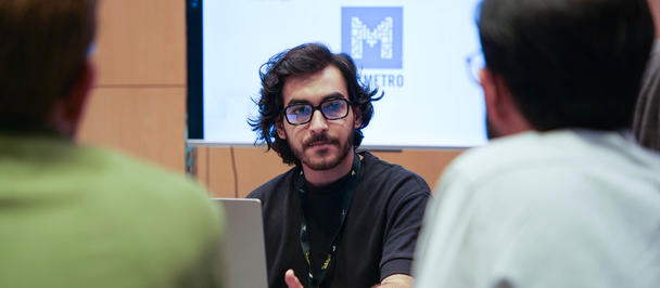 Man with glasses talks to two attendees in a conference room; presentation slide in the background.