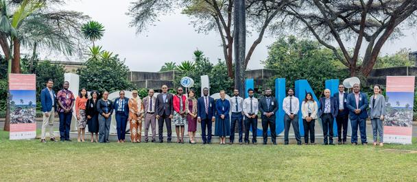 Group photo: diverse people lined up outdoors in a park with banners and trees in the background.