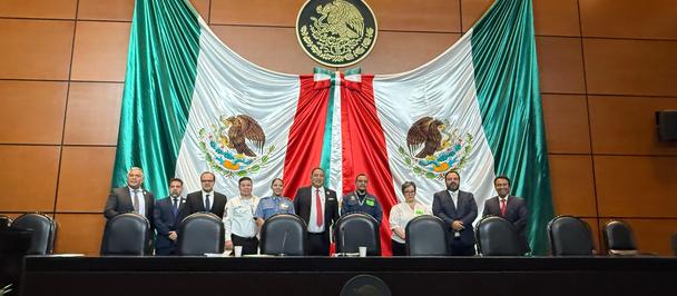 Panel of officials on a stage behind a long desk, framed by large Mexican flags.