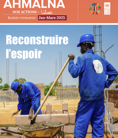Two workers in blue coveralls and helmets lay bricks at a site beneath an orange banner.