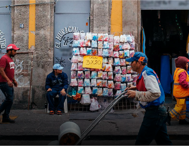 Men walk past a vendor selling colorful bags, with a bright yellow wall behind them.