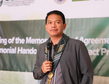 Speaker in dark ceremonial robe with a gold medallion, holding a microphone on a ceremony stage.