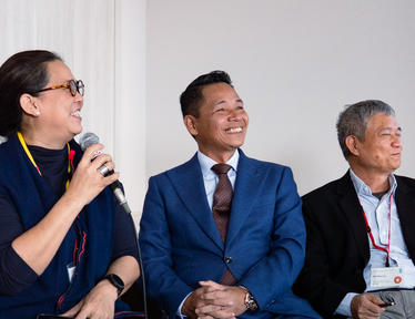Panel of three professionals in a bright room; left speaker holds a microphone while others listen.