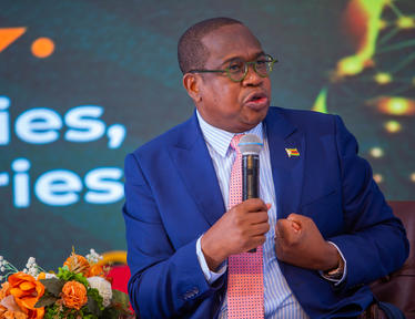 Man in blue suit and pink tie speaks into a microphone on stage; orange flowers nearby.