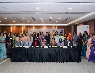 Diverse group in formal attire posing behind a long table draped with a black cloth on a stage in a conference room.