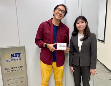 Two people in a hallway; one in red shirt and yellow pants holds a card, KIT sign left.