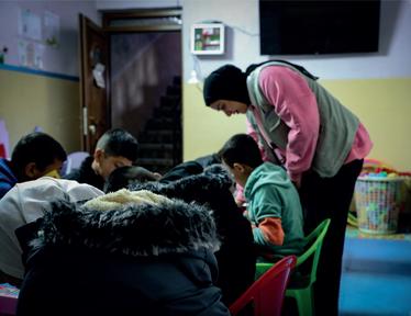 Caregiver in pink shirt and green vest helps children at a table in a classroom.