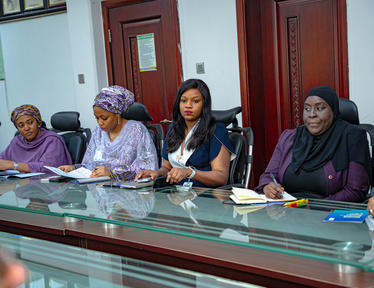 Five women, some wearing hijabs, sit around a glass conference table with papers.