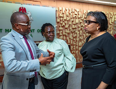 Man in suit hands something to green-clad woman as another woman watches.
